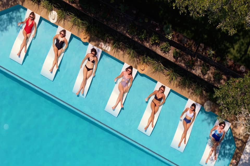 Overhead view of resort pool with guests on white lounge loungers, turquoise water, and palm tree shadows