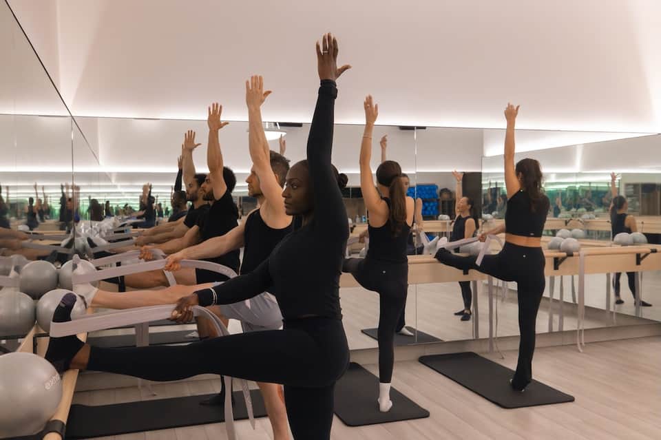 Group fitness class doing Pilates reformer exercises with mirrors and city views in background