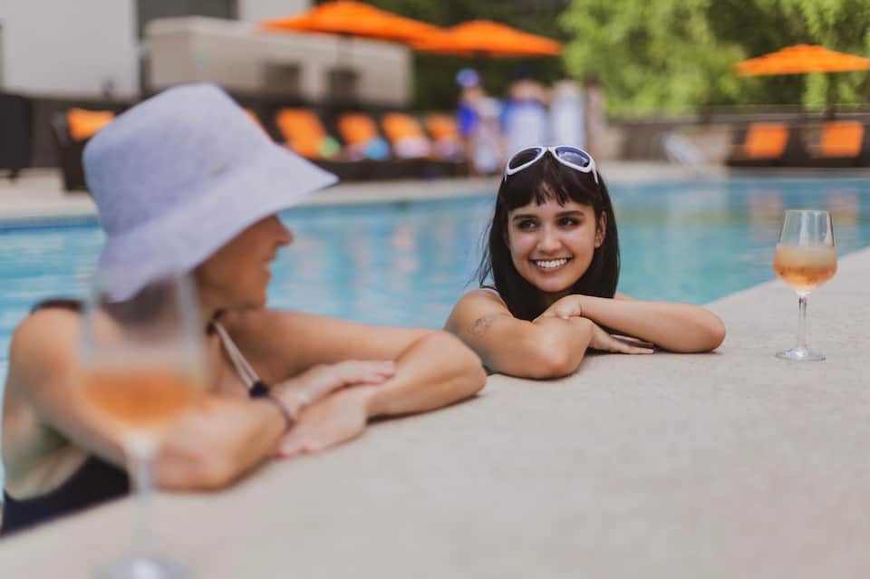 Woman at pool edge with drink, white sun hat visible, blue pool water in background