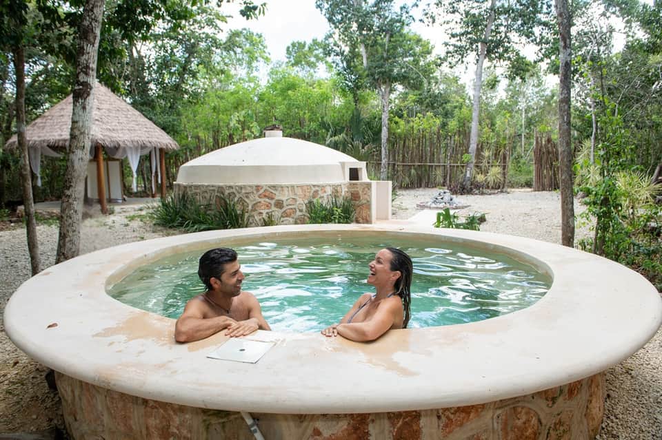 Couple relaxing in circular outdoor hot tub surrounded by forest trees and tiki huts