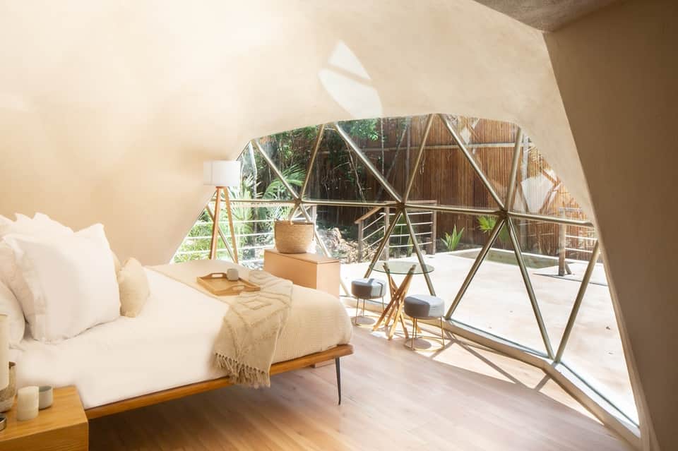 Luxury glamping bedroom with white linens, wooden platform bed, and geometric glass-walled outdoor patio