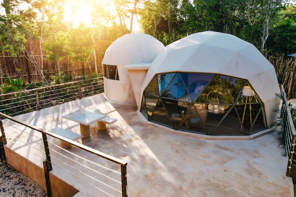 Two white dome-shaped glamping structures with geometric windows, wooden deck, and forest setting