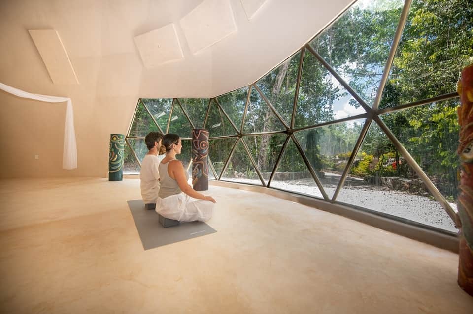Two people meditating in modern geodesic dome with triangular windows overlooking nature trail