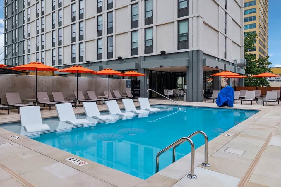 Urban hotel rooftop pool with white loungers, orange umbrellas, and city skyline backdrop