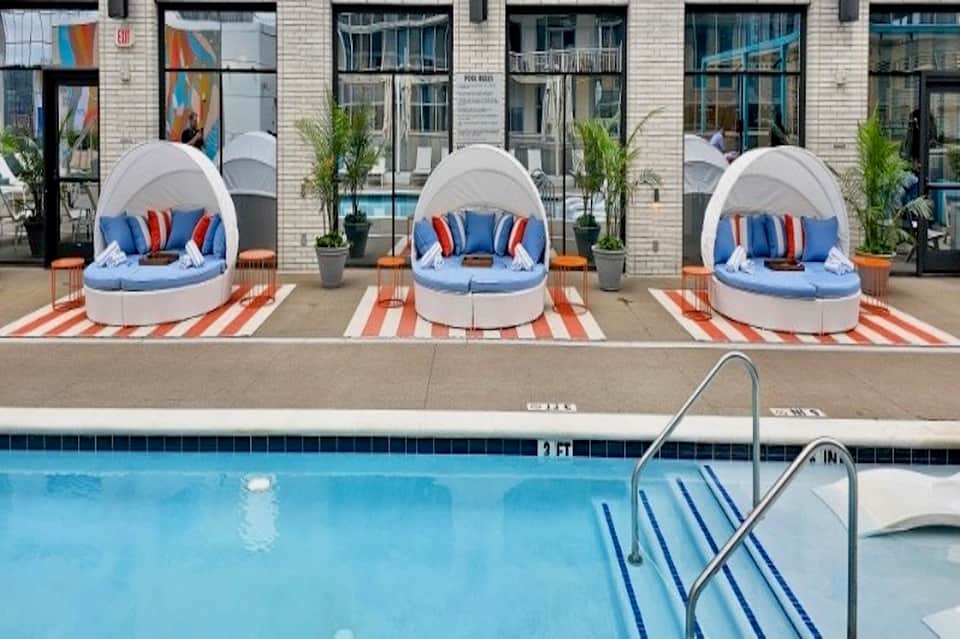Three white pod-shaped cabanas with blue cushions on striped patio beside hotel pool and brick building