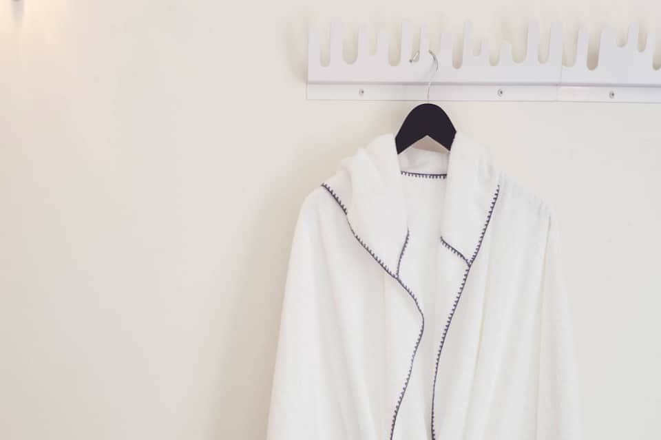 White spa robe with blue stitching hanging on black hanger against white wall