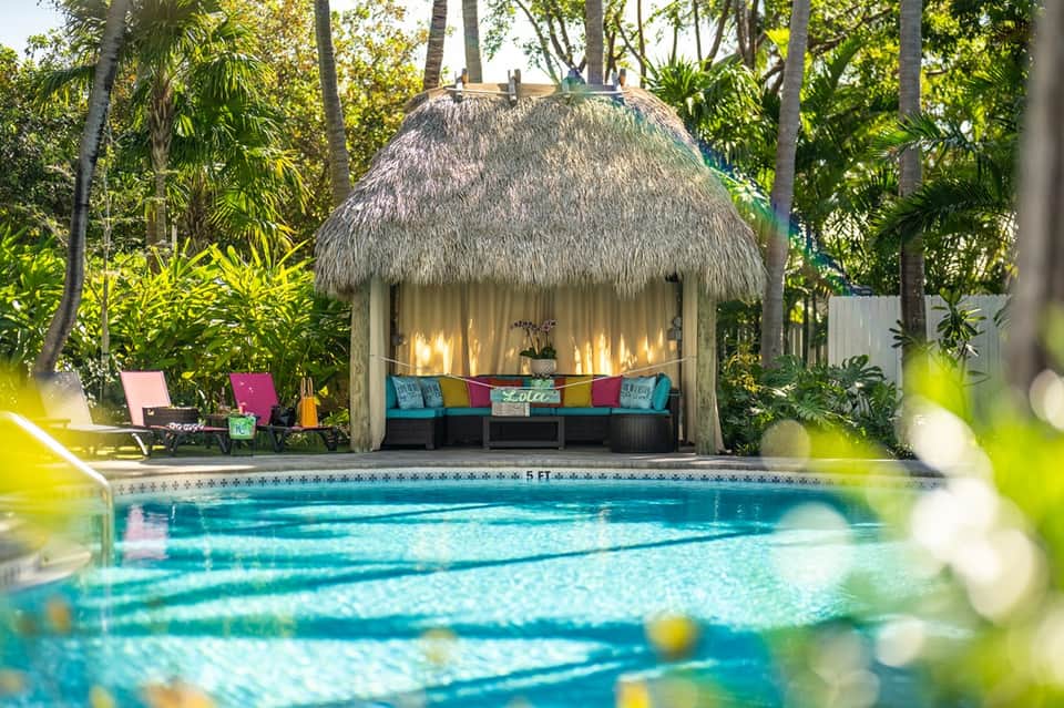 Lap pool with thatched-roof cabana, colorful pillows, tropical trees, lounge chairs, and lush landscaping