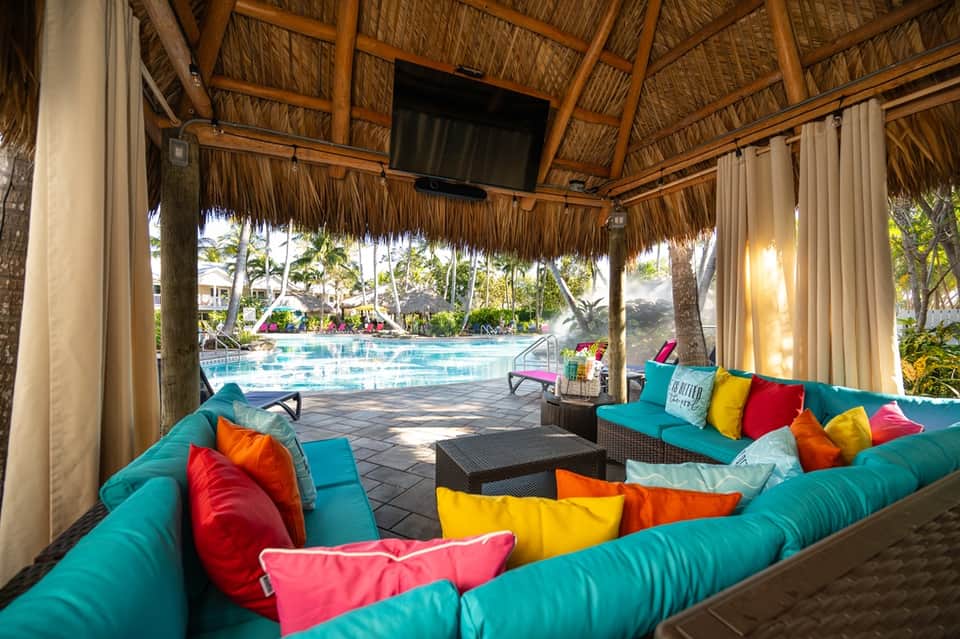 Thatched-roof pool pavilion with turquoise seating, colorful pillows, and view of resort pool beyond