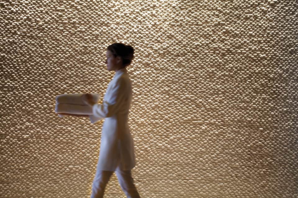 Spa attendant walking past textured beige wall in wellness facility wearing white robe
