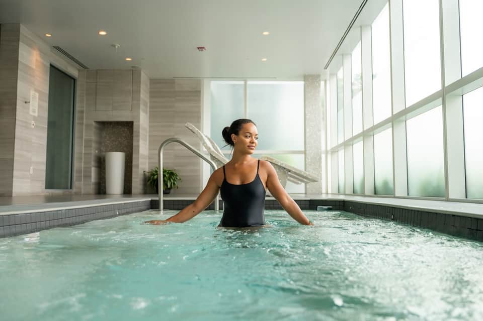 Woman relaxing in modern spa tub with floor-to-ceiling windows and neutral tile design