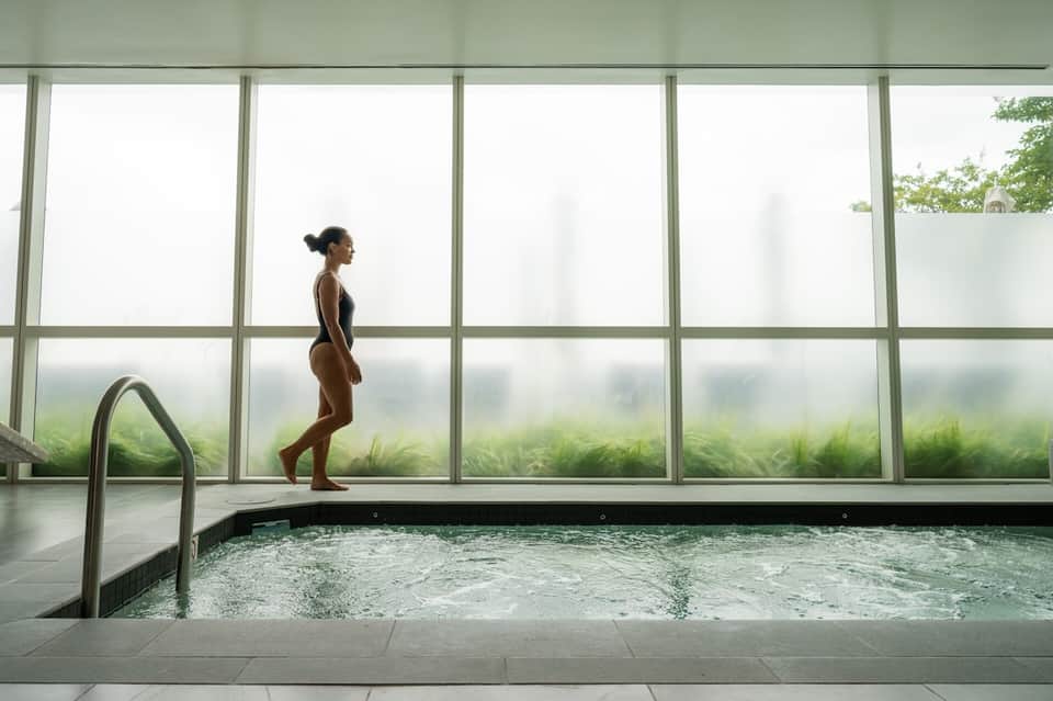 Woman standing by indoor spa pool with large windows overlooking green landscape and trees