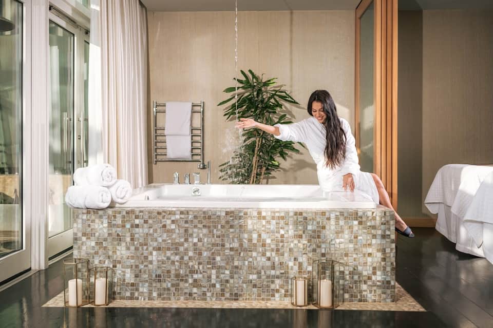 Woman in white robe arranging flowers beside luxurious bathroom tub with tile surround