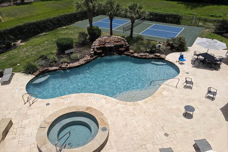 Resort pool with waterfall feature, hot tub, tennis courts, and palm trees in background