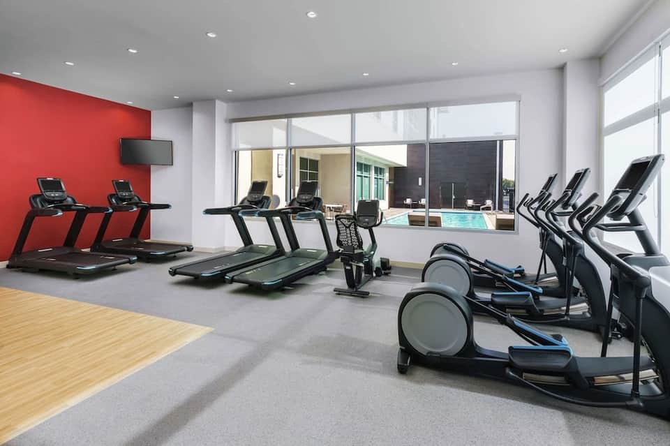Bright fitness center with treadmills, exercise bikes, red accent wall, and pool view through large windows