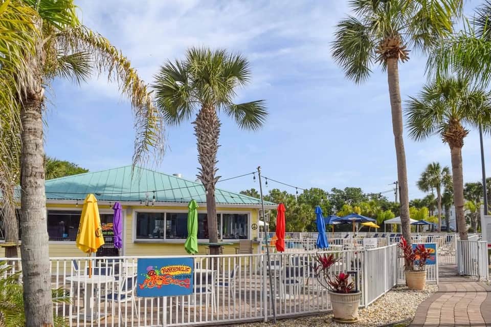Beachfront dining area with colorful umbrellas, palm trees, and casual restaurant with green roof under cloudy sky