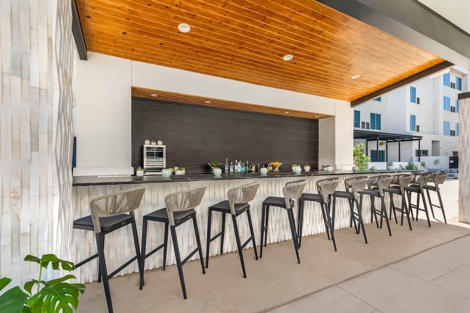 Outdoor bar with wooden ceiling and dark backsplash, featuring woven bar stools and modern design