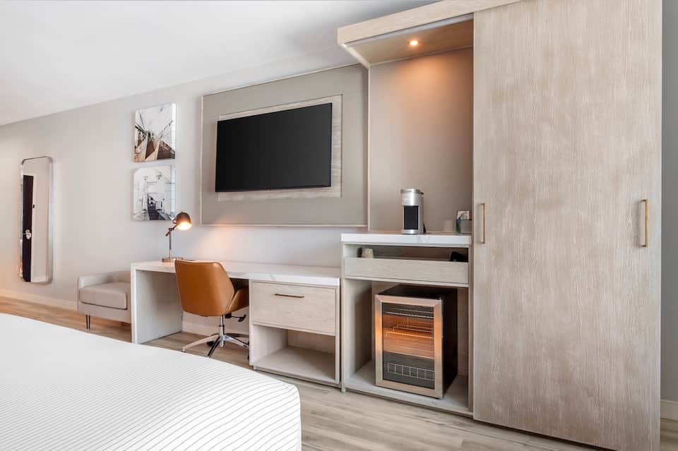Modern hotel room with work desk, flat-screen TV, coffee station, wardrobe, and contemporary furnishings