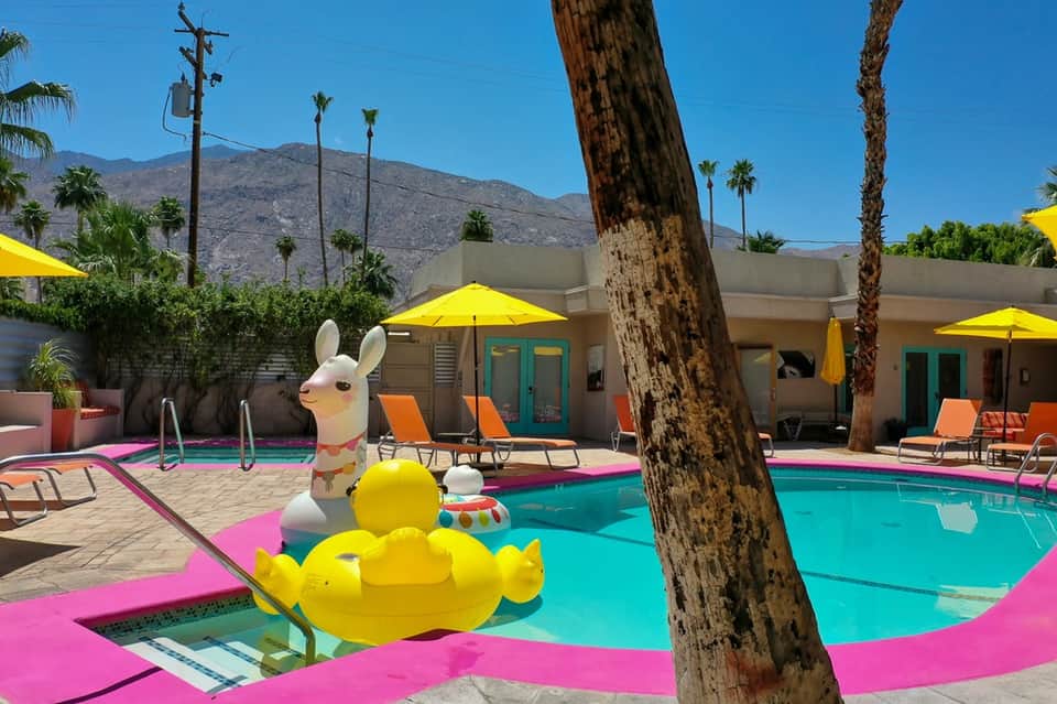 Colorful resort pool with pink decking, yellow umbrellas, playful pool floats, and mountain views
