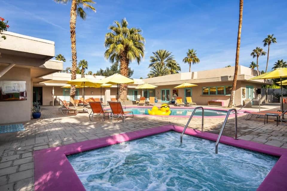 Resort courtyard with purple hot tub, yellow umbrellas, palm trees, and colorful loungers
