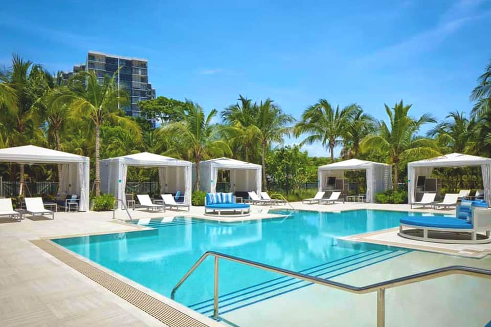 Luxury resort pool with white cabanas, palm trees, and high-rise hotel building under clear blue sky