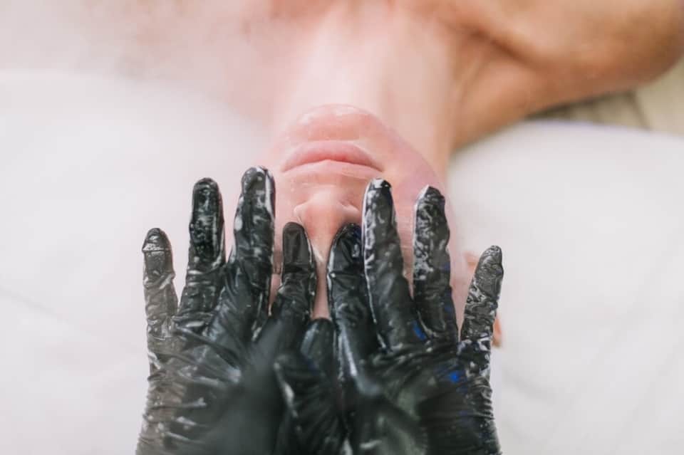 Close-up of hands covered in black spa mud or treatment substance against white towel background.