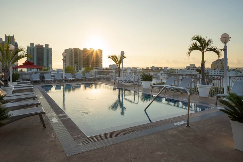 Rooftop pool at sunrise with cityscape, palm trees, and lounge chairs