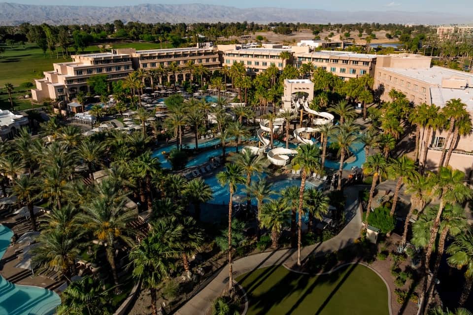 Aerial view of large resort complex with multiple pools, palm trees, and desert mountain backdrop