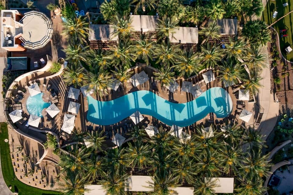 Aerial view of resort with turquoise wave-shaped pool, palm trees, white loungers, and circular building structures