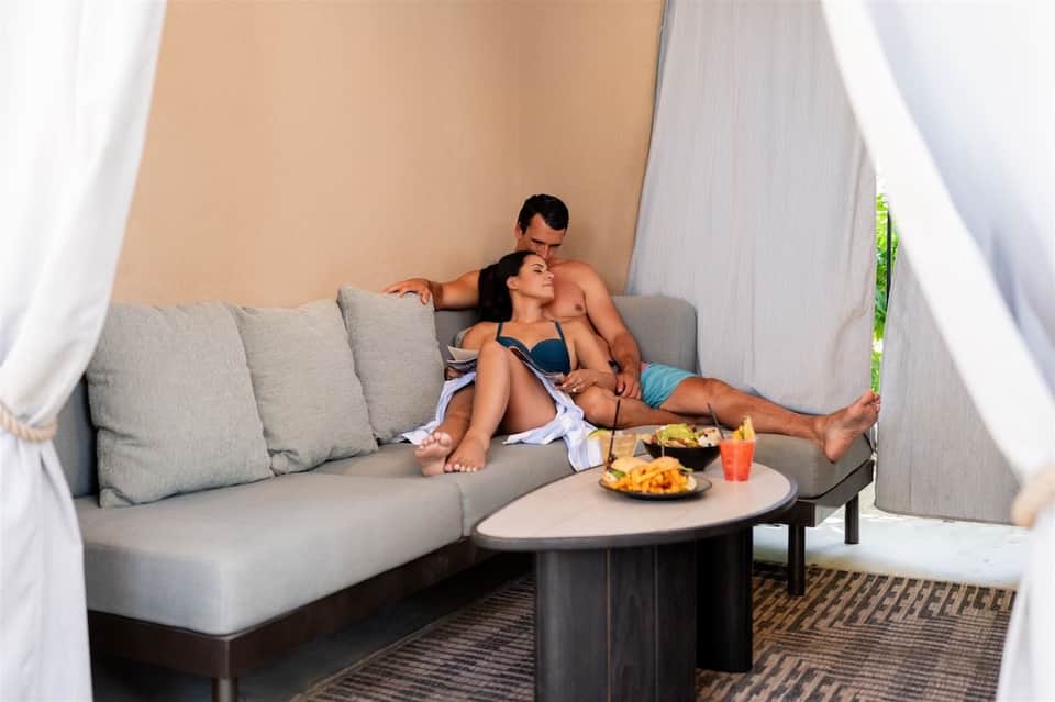 Couple relaxing on outdoor daybed with white curtains, food and drinks on side table