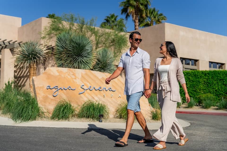 Couple walking past Agua Serena Spa entrance sign on sunny grounds with desert vegetation