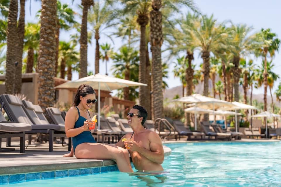 Couple enjoying drinks at pool edge with palm trees, lounge chairs, and umbrellas in background
