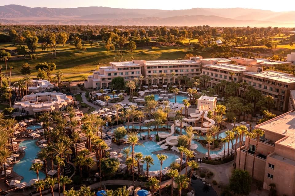 Aerial view of resort with multiple pools, palm trees, and pink buildings in desert valley landscape