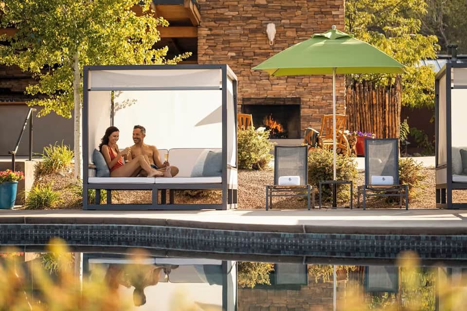 Couple relaxing in canopied daybed lounge near pool with green umbrella and brick fireplace backdrop