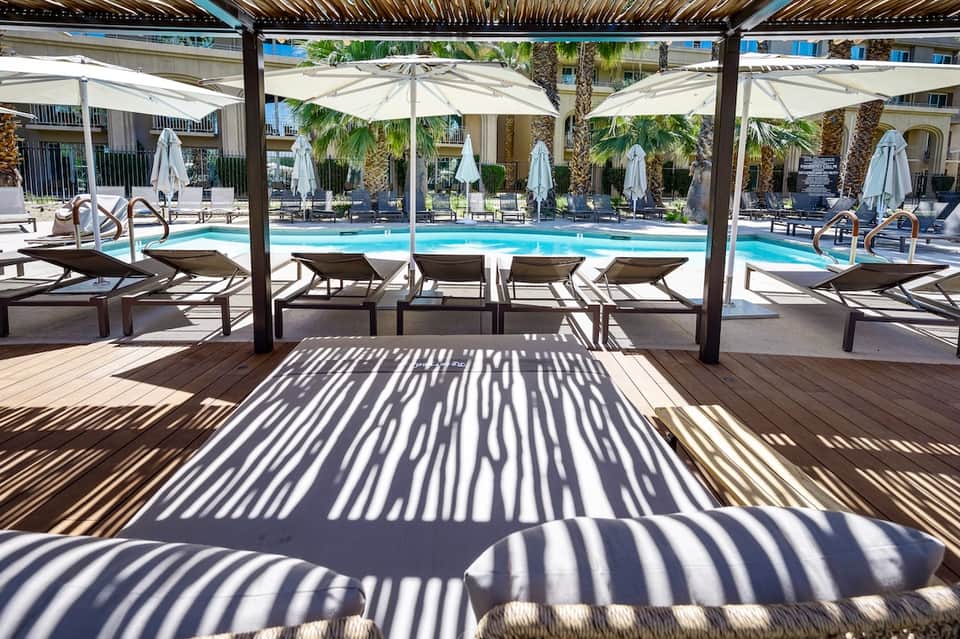 Resort pool deck with lounge chairs under pergola shade, white umbrellas, and palm trees with bright blue pool water