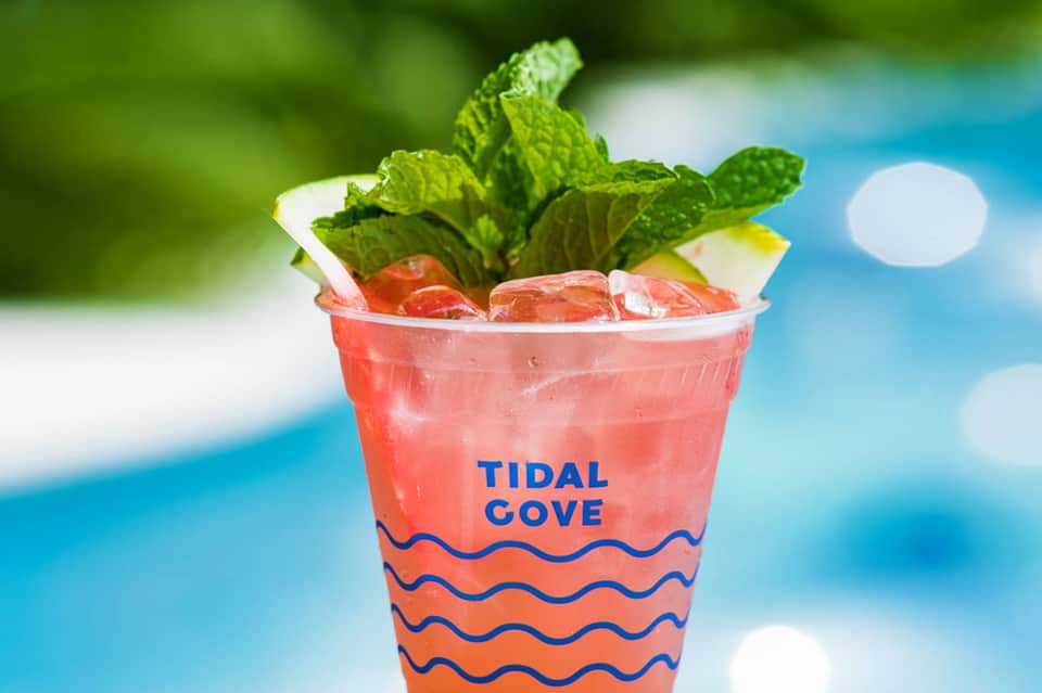 Refreshing coral-colored cocktail with mint garnish in Tidal Cove branded cup against blurred blue water background