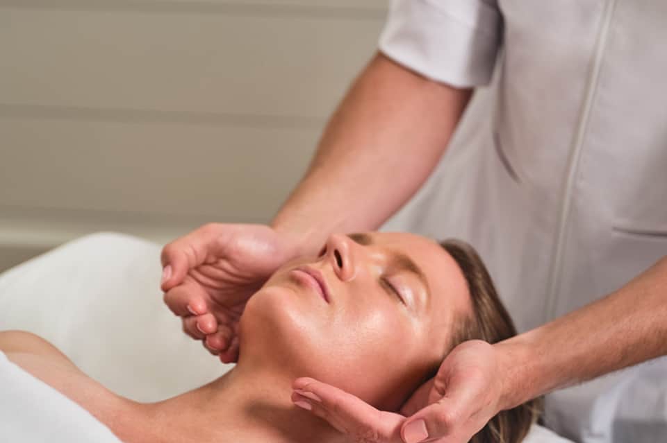 Spa therapist performing facial massage on relaxed woman in white treatment room