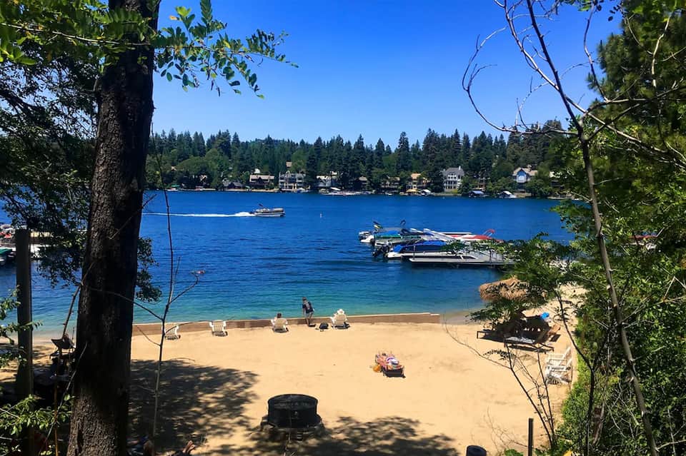 Sandy beach with boats, dock, clear blue water, and tree-lined shoreline with residential homes