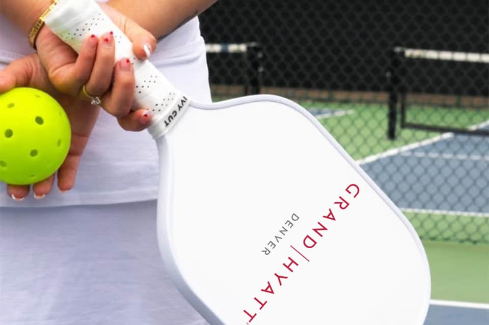 Close-up of woman's hand holding pickleball paddle with bright yellow ball on indoor court with net visible
