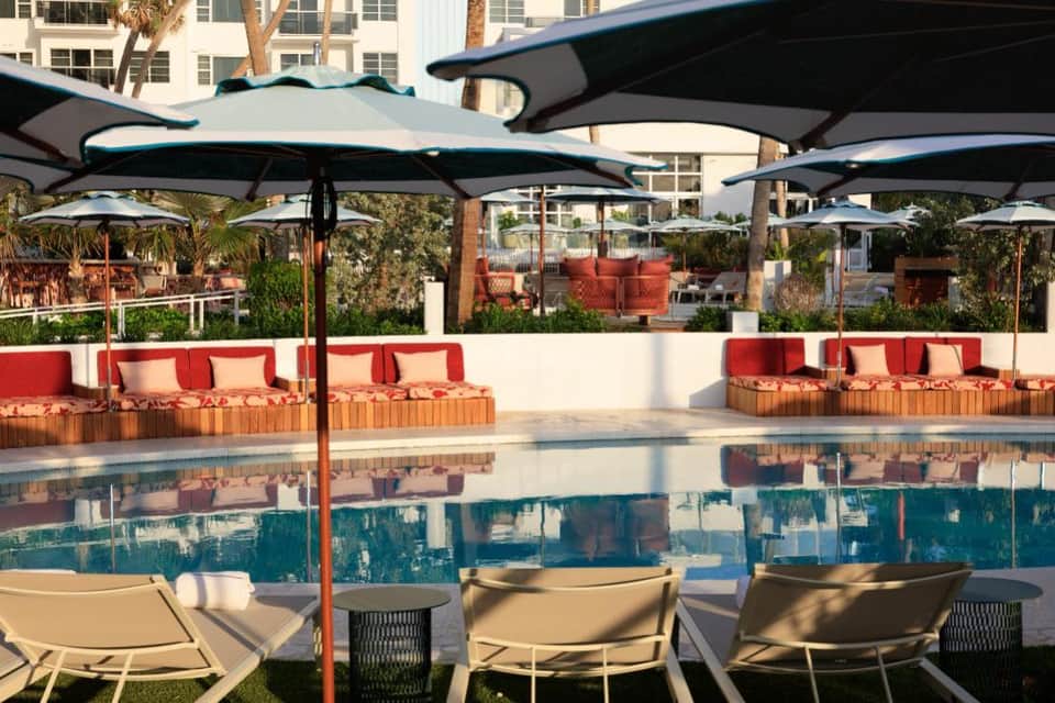 Resort pool deck with turquoise water, red lounge seating, teal umbrellas, palm trees, and high-rise buildings