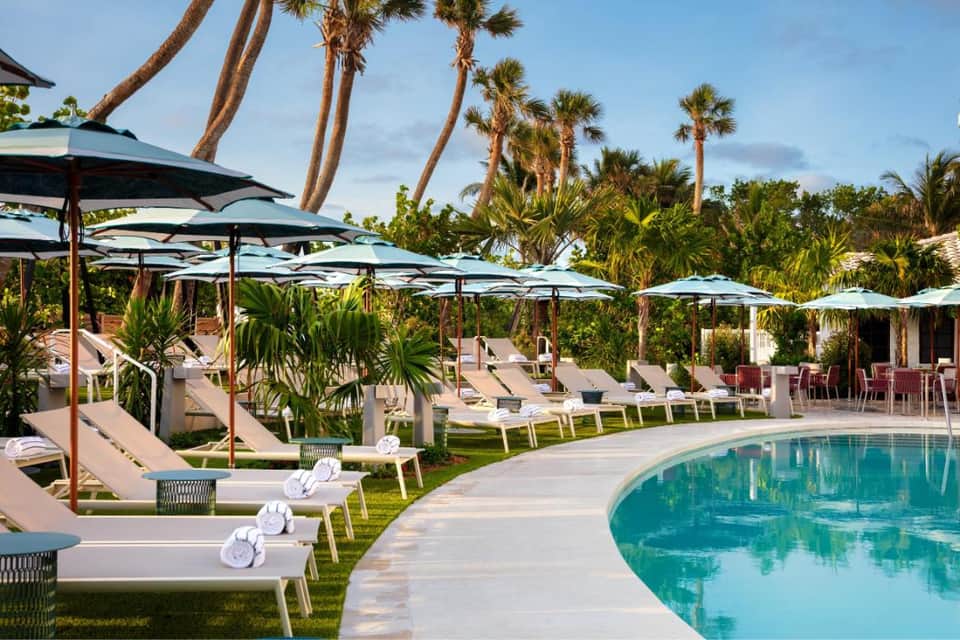 Resort pool area with white loungers, dark umbrellas, palm trees, and lush landscaping