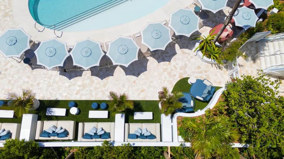 Aerial view of beachfront pool with blue umbrellas and cabanas on white sand near water