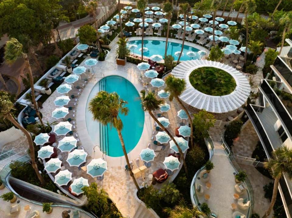 Aerial view of resort pool complex with light blue umbrellas, palm trees, and surrounding buildings