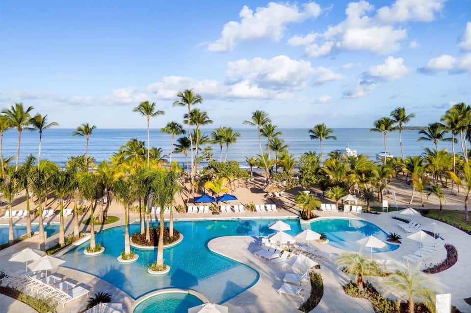 Aerial view of luxury resort pool with palm trees, white umbrellas, loungers, and ocean view beyond