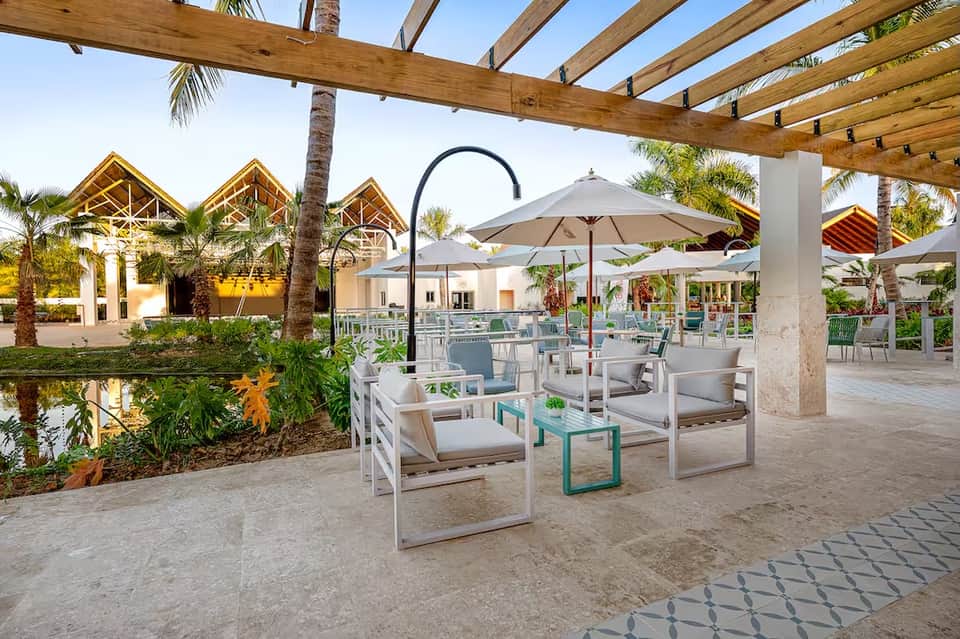 Resort courtyard with white and teal furniture, umbrellas, pergola, palm trees, and water feature