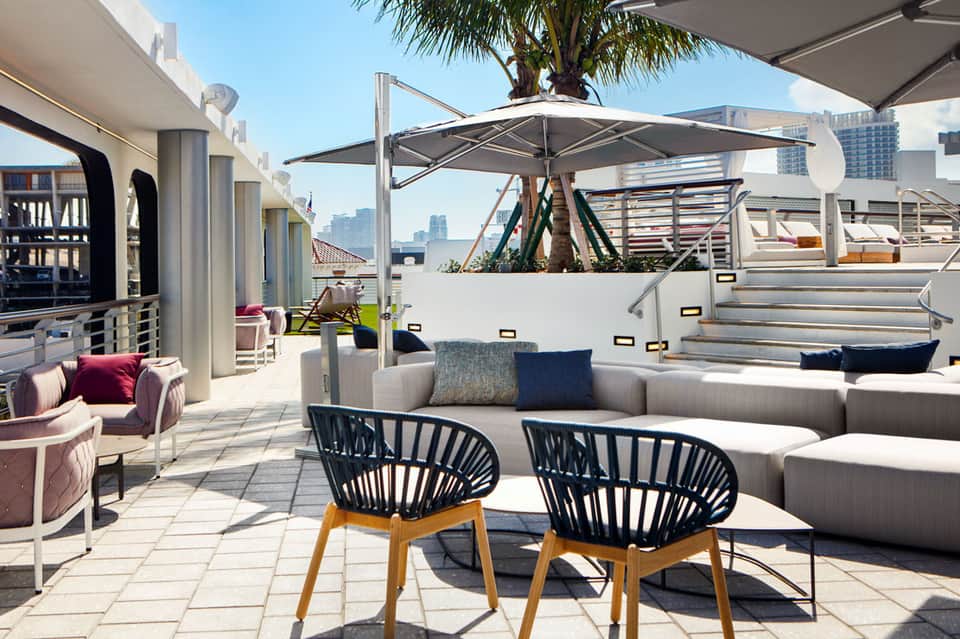 Modern rooftop lounge with contemporary furniture, umbrellas, palm trees, and city skyline views
