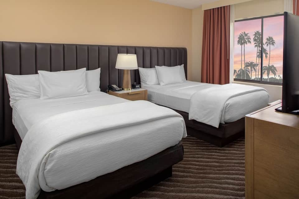 Hotel guest room with two queen beds, gray headboards, warm lighting, and tropical sunset view