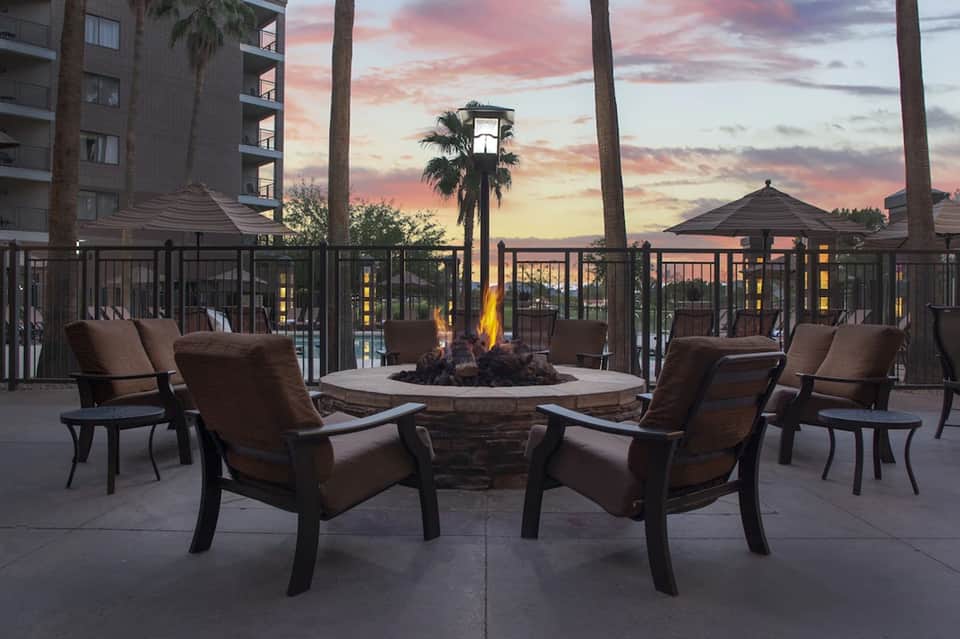 Outdoor fire pit lounge at dusk with brown cushioned chairs, palm trees, and sunset sky with building in background.