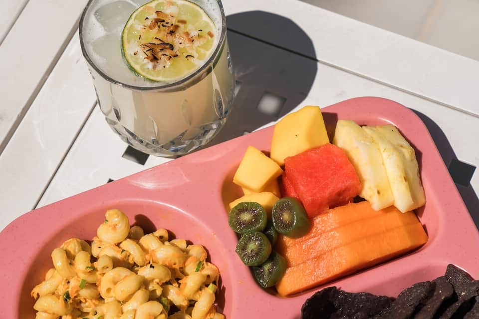 Pink plate with fresh fruit and pasta dish, cocktail with lime garnish on white table