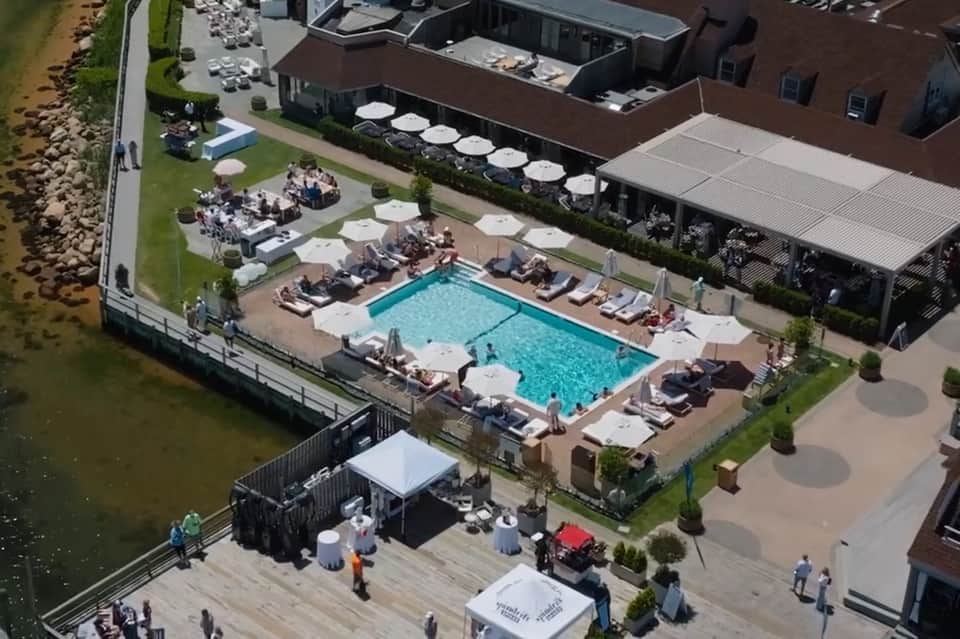 Aerial view of waterfront resort pool with lounge chairs, umbrellas, adjacent building, and event setup
