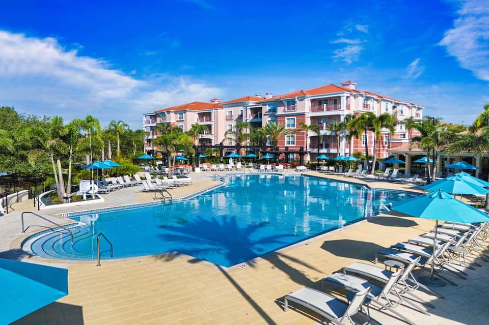 Large resort pool with pink building, blue umbrellas, lounge chairs, and palm trees
